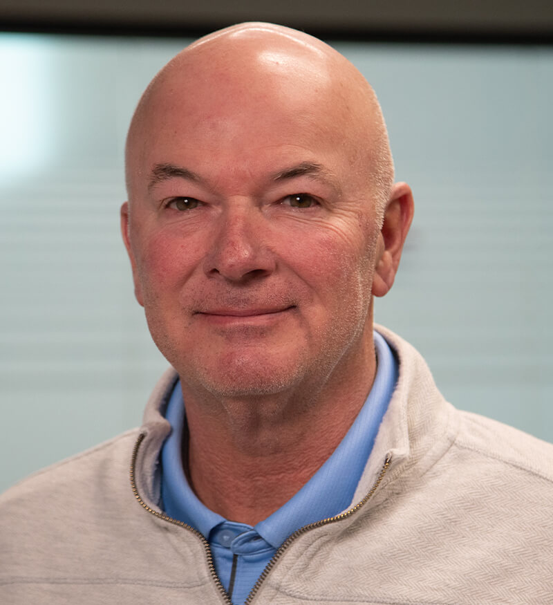 A middle-aged man with a bald head is wearing a light gray zip-up sweater over a blue collared shirt, standing in front of a blurred background. He is looking at the camera and smiling slightly.