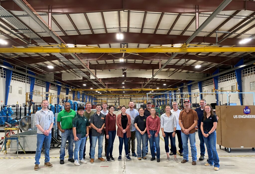 A group of 18 people stands in a single row inside a large industrial warehouse, facing the camera. The background shows machinery, equipment, and high warehouse ceilings.