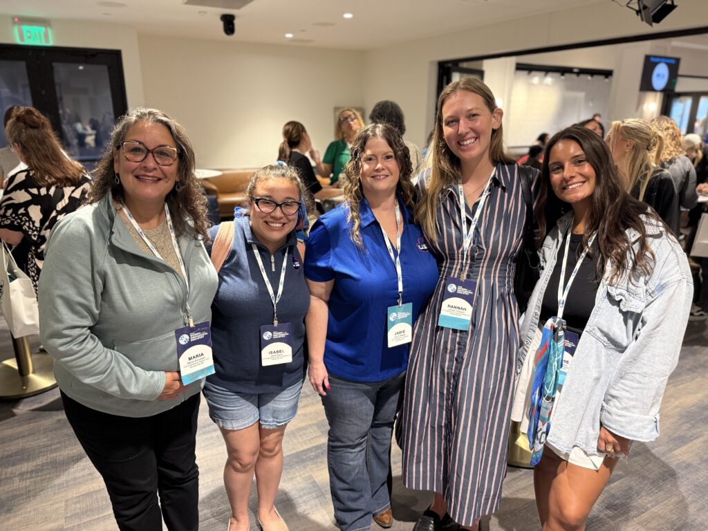 Five women stand side by side smiling at a conference, each wearing name badges and casual to business casual clothing. Other attendees are visible in the background of the indoor event space.