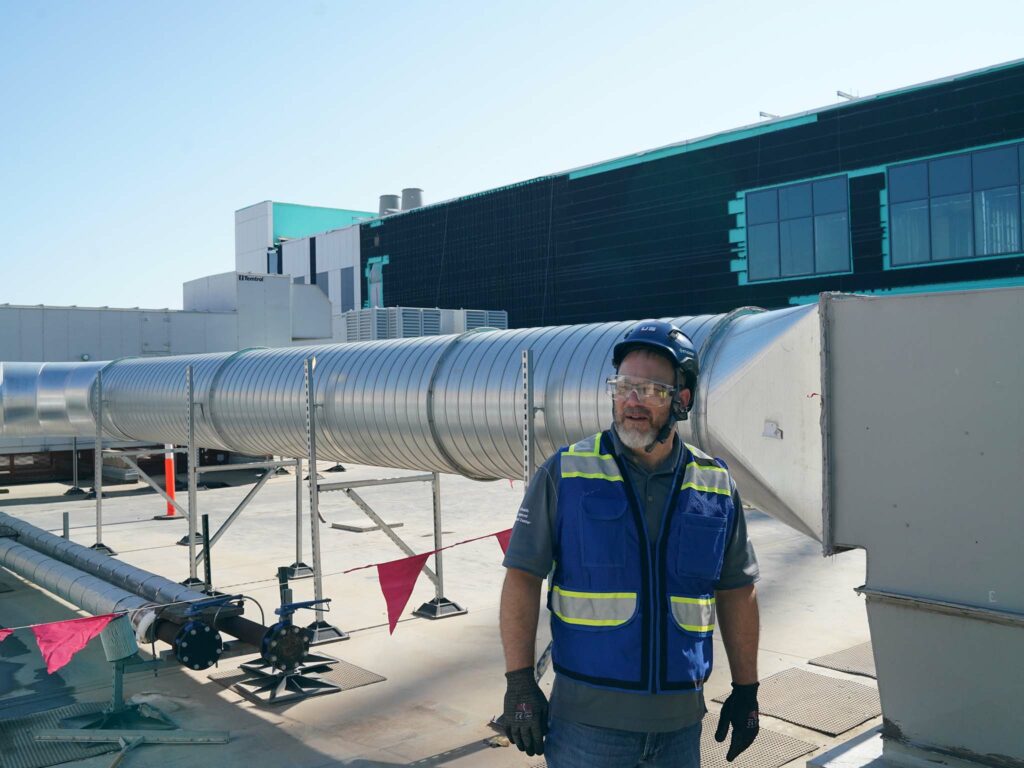 A man wearing a blue safety vest, helmet, gloves, and protective glasses stands on a rooftop near large industrial ventilation ducts, overseeing construction activities. A modern building with teal accents is visible in the background.