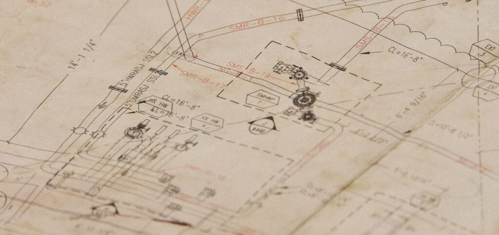 A close-up of a technical engineering or architectural blueprint with various lines, symbols, numbers, and labels in black and red ink on slightly worn paper.