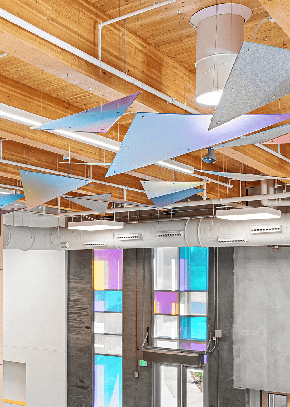 A modern interior space with a wooden ceiling, exposed ducts, geometric metal panels hanging from the ceiling, and tall windows with panels of colored glass letting in natural light.