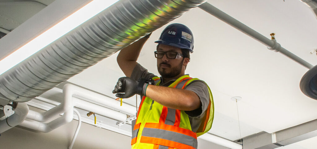 A worker wearing a blue hard hat, safety glasses, gloves, and a reflective vest adjusts or inspects a large metal air duct on a ceiling with exposed pipes and lights.