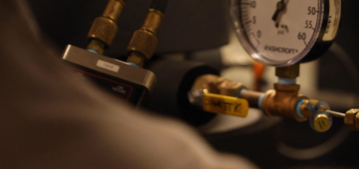 Close-up view of industrial equipment featuring brass fittings, a pressure gauge, and a yellow-handled valve. The gauge shows a scale from 0 to 100, with some elements out of focus in the foreground.
