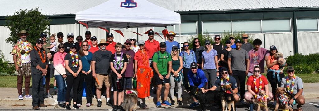 A group of people, some wearing leis and tropical shirts, pose outdoors in front of a building and a white tent. Several dogs are present, and the group appears to be gathered for a casual event.