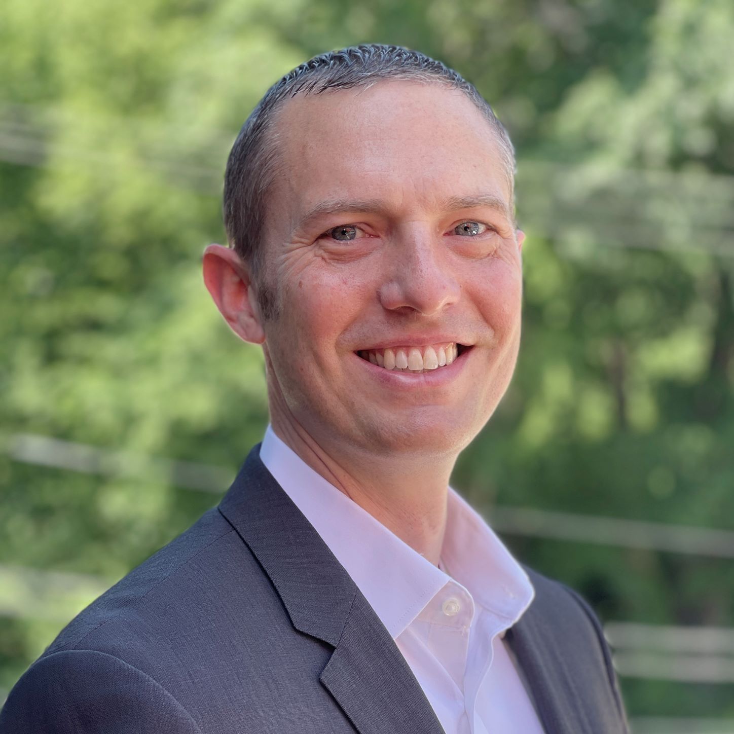 A man in a dark suit and white shirt smiles at the camera, standing outdoors with green trees and blurred power lines in the background.