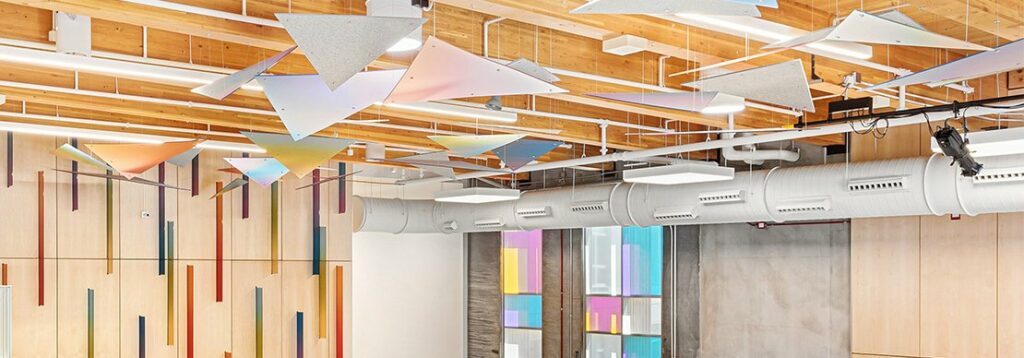 A modern interior space with a wooden ceiling, exposed ductwork, and triangular acoustic panels. Colorful rectangular accents are on the wall and windows, and the lighting is bright and even.