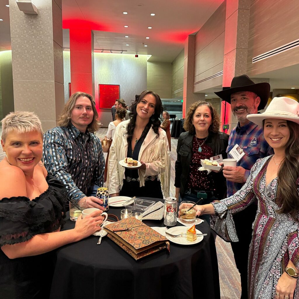 Six adults, three men and three women, stand around a high-top table at an indoor event. They are dressed in semi-formal or western-style attire, smiling and holding drinks and plates of food.