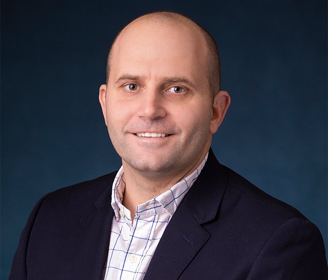 A man with short hair smiles at the camera. He is wearing a dark blazer over a white, checkered shirt. The background is a solid dark blue.