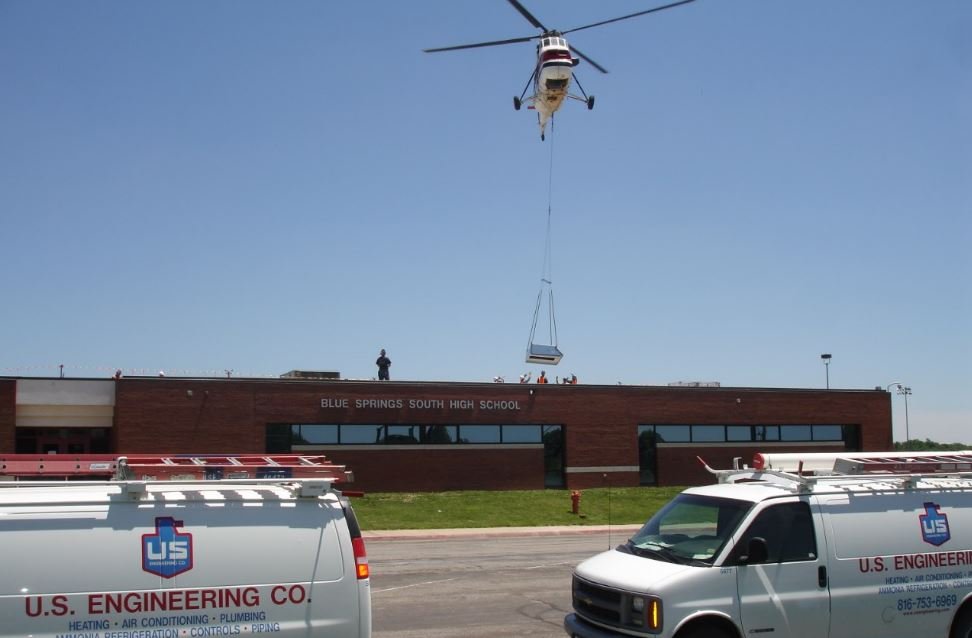 A helicopter lowers equipment onto the roof of Blue Springs South High School while two U.S. Engineering Co. vans are parked in the foreground. The weather is clear and sunny.