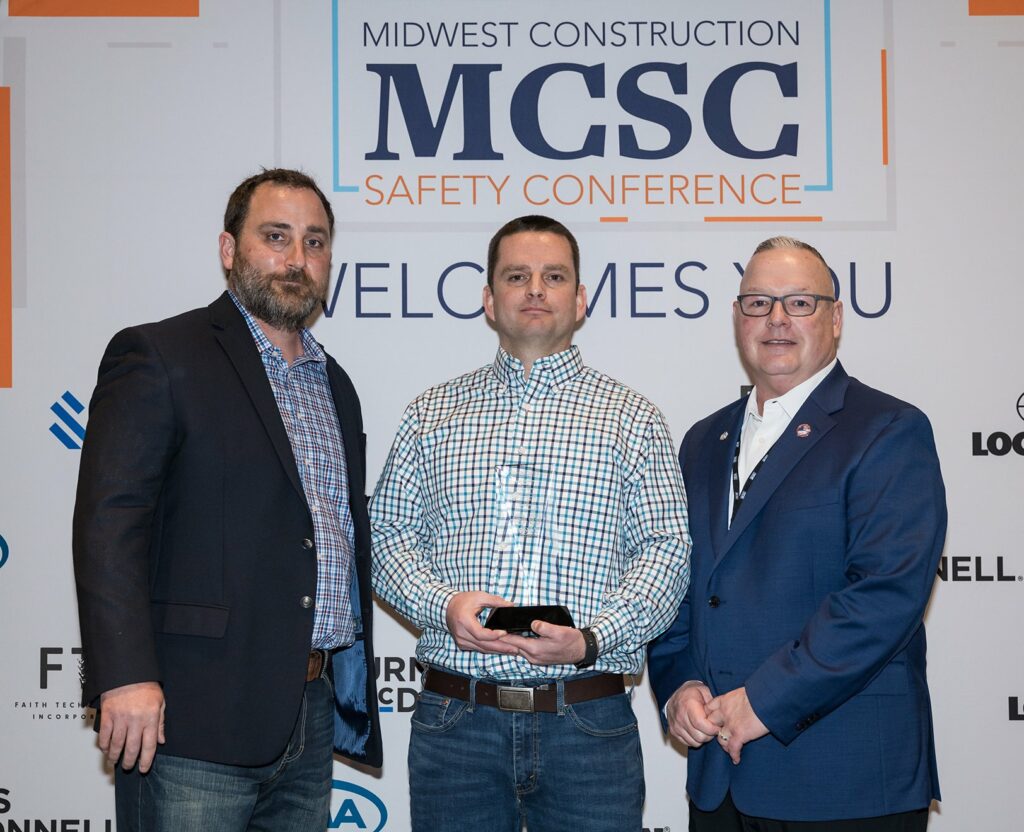 Three men stand together in front of a Midwest Construction Safety Conference backdrop. The man in the center is holding a black object, while the men on either side are standing with their arms at their sides.