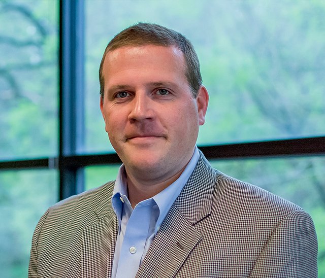 A man in a light blue dress shirt and checkered blazer is standing indoors in front of large windows with a view of trees and greenery outside.