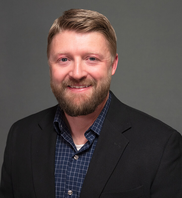A man with light brown hair and a beard, wearing a black blazer over a blue checkered shirt, is smiling in front of a plain gray background.