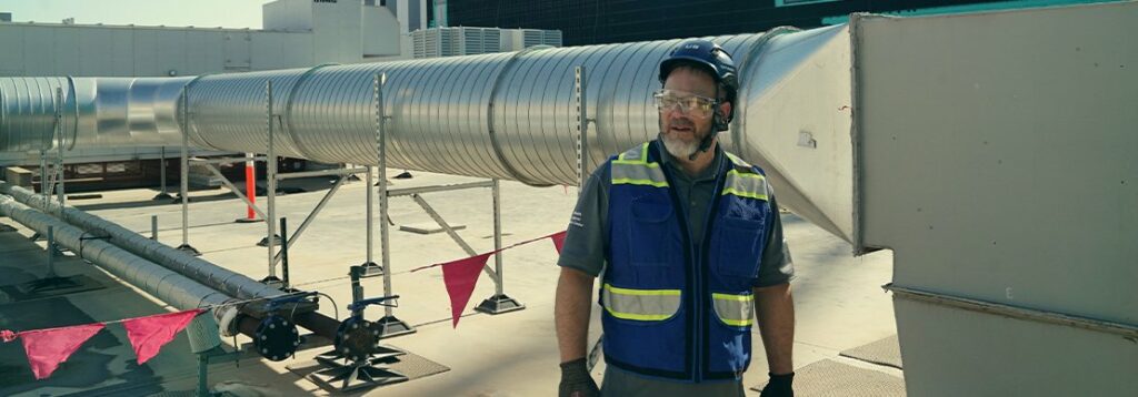 A worker wearing a blue safety vest, gloves, helmet, and safety goggles stands on a rooftop near large metal ventilation ducts and red warning flags. Industrial equipment is visible in the background.