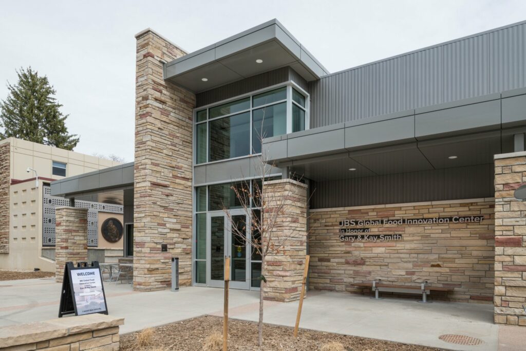 The exterior of the JBS Global Food Innovation Center at Colorado State University, featuring a modern building with stone and metal accents, large windows, and a welcome sign on a stand near the entrance.