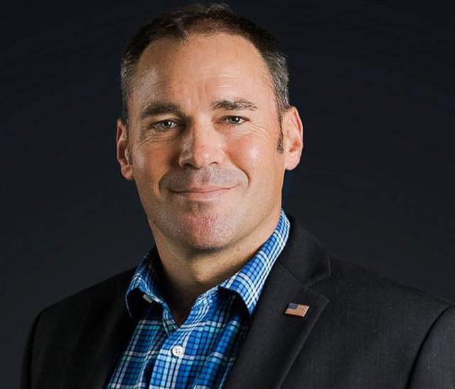 A man in a dark suit jacket and blue plaid shirt, wearing an American flag pin, stands against a dark background, looking at the camera with a slight smile.