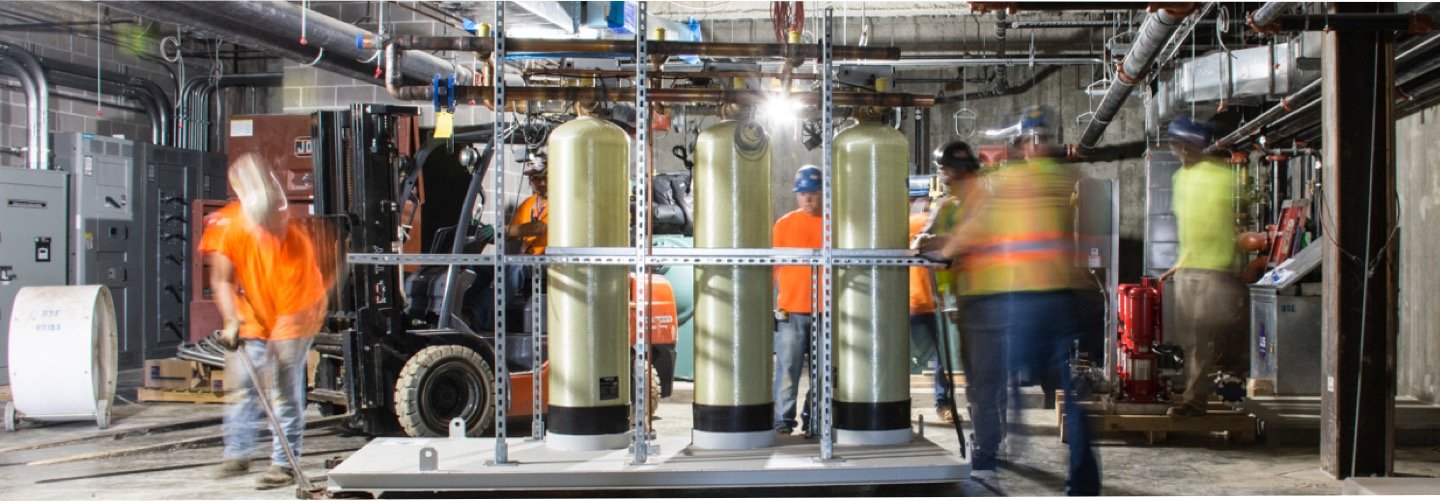 Construction workers in safety gear work around large cylindrical tanks and various machinery in an industrial indoor setting with visible pipes, wires, and equipment. Some workers appear blurred, indicating movement.