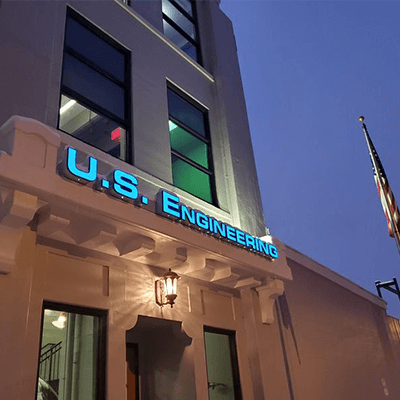 A building exterior at dusk with a lit-up sign that reads U.S. ENGINEERING. An American flag is visible on the right side of the image. Windows and a lit entrance door are also shown.