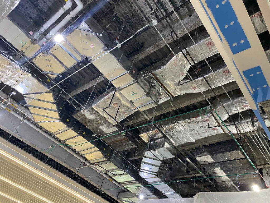 Exposed ceiling showing HVAC ducts, insulation, wiring, and lighting fixtures in a commercial building under construction. Some sections are unfinished, with visible pipes and building materials, highlighting ongoing construction and potential offsite fabrication work.
