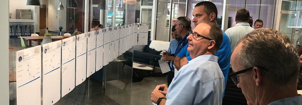 Several men stand in an office, closely examining a glass wall covered with large sheets of paper containing charts or schedules. The setting appears collaborative and professional.