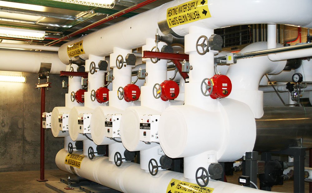 A row of large insulated pipes with valve wheels and red control units, labeled with yellow caution signs, inside an industrial mechanical room with exposed ducts and a concrete wall.