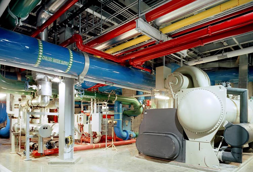 Industrial mechanical room with large blue, red, and green pipes on the ceiling and walls, labeled “chilled water supply,” alongside various machinery, valves, and electrical equipment on a polished concrete floor.