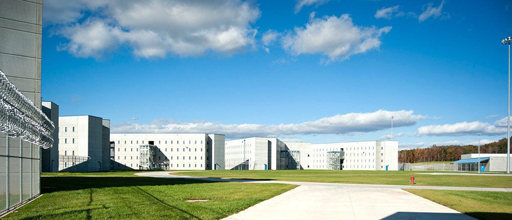 A large, modern correctional facility with multiple gray buildings, tall windows, and metal staircases, surrounded by green grass, fencing, and razor wire under a blue sky with scattered clouds.