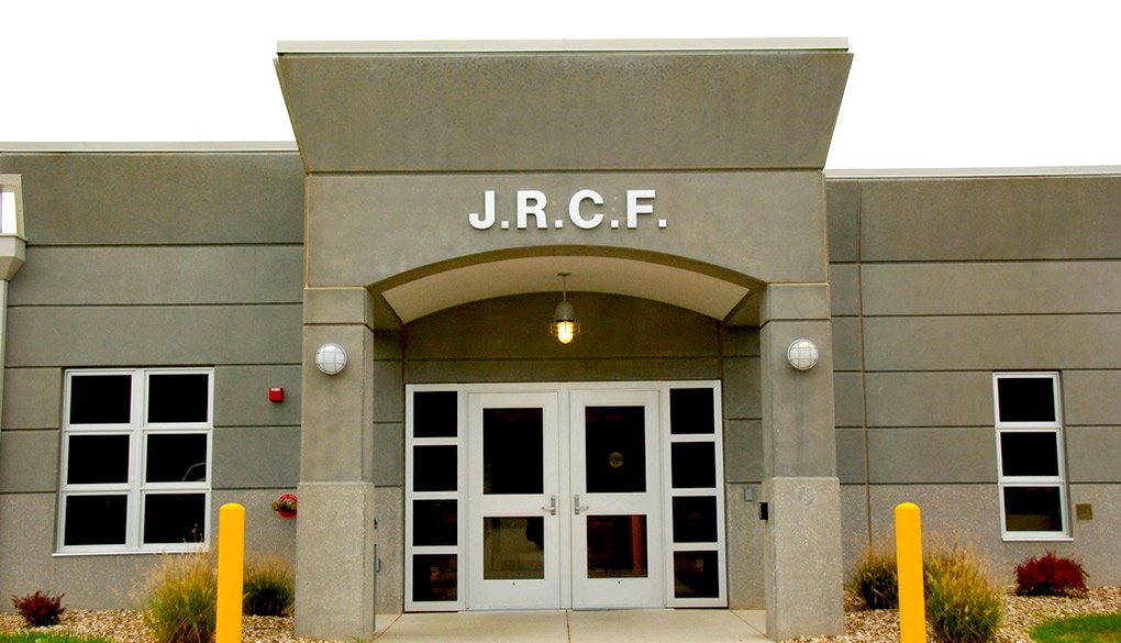 The front entrance of a gray building labeled J.R.C.F. above double glass doors, with small windows on each side, exterior lights, and a few yellow posts and plants in front.