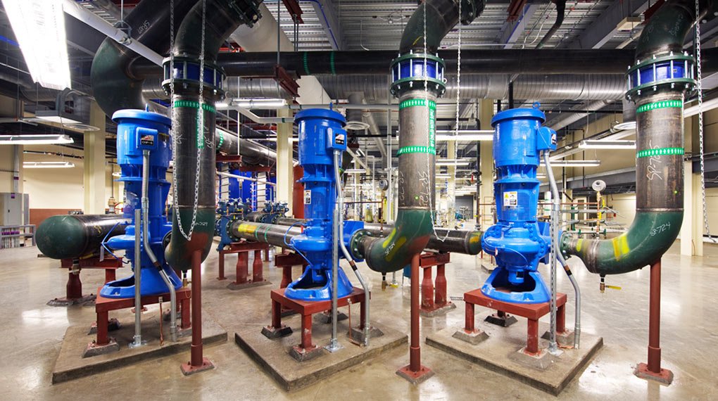 Large industrial room with five blue industrial pumps connected to green and black piping, mounted on red metal supports, with exposed ceilings and concrete floor.