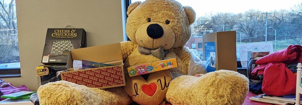 A large teddy bear sits on a table holding a heart-shaped pillow and surrounded by board games, a shoe box, and various items. A window in the background shows trees and a cloudy sky.