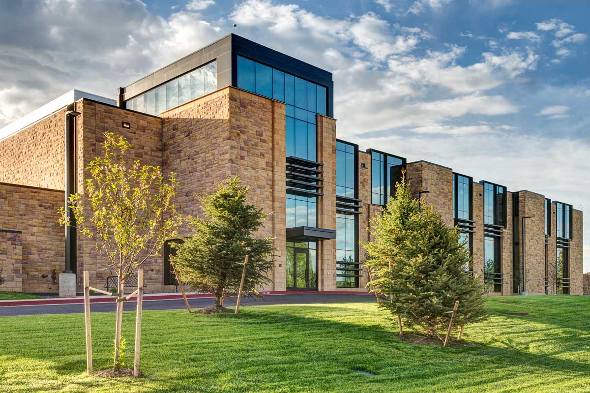 A modern building with large glass windows and tan stone walls stands on a well-manicured lawn with several small trees under a partly cloudy sky.