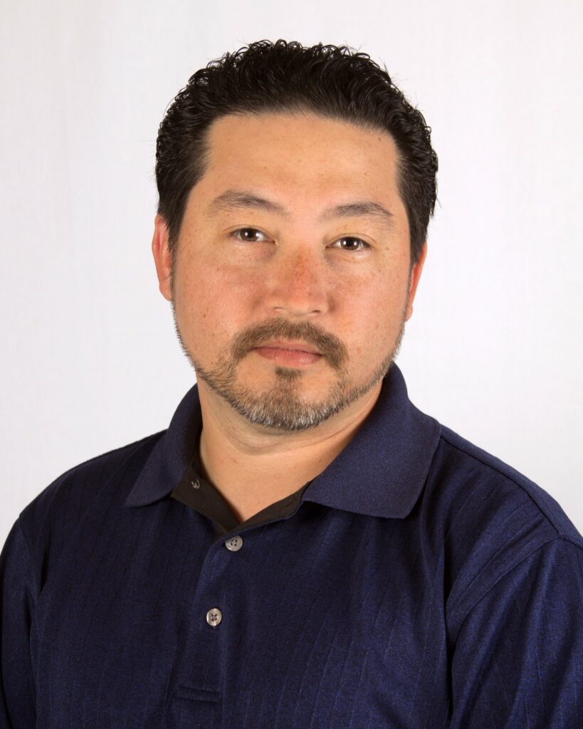 A man with short dark hair, a trimmed beard, and mustache is wearing a dark blue collared shirt. He is facing the camera with a neutral expression against a plain, light-colored background.
