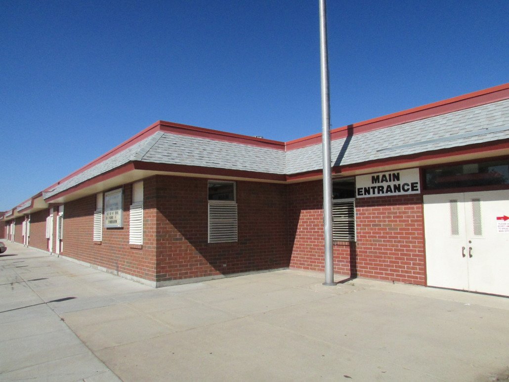 A one-story brick building with a gray roof and red trim. A sign above double doors reads Main Entrance. The sidewalk and a tall flagpole are visible in front of the building under a clear blue sky.