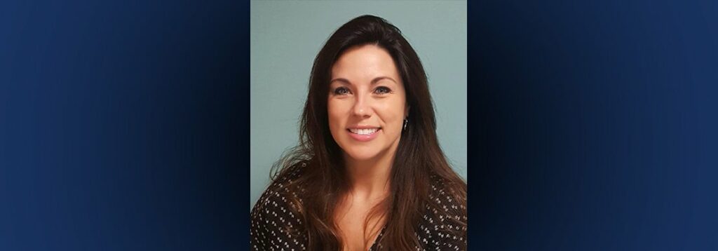 A woman with long brown hair, wearing a black top with white polka dots, smiles at the camera in front of a plain light blue background.