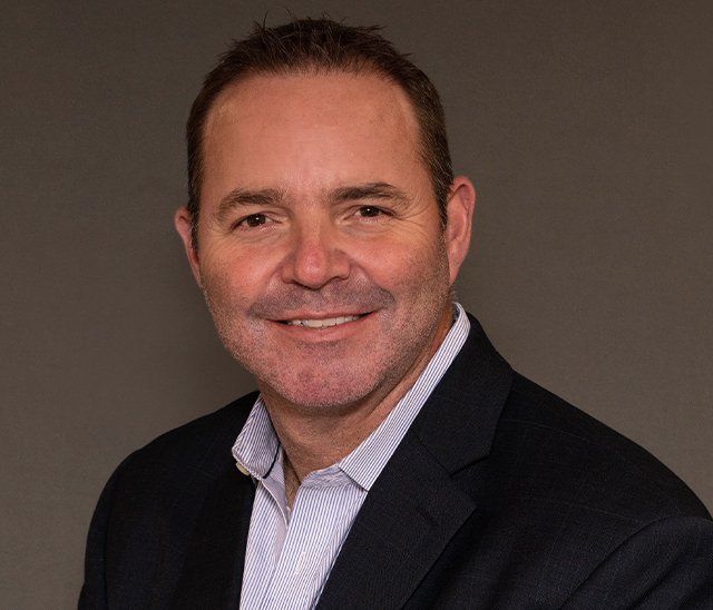 A man with short brown hair, stubble, and wearing a dark suit jacket over a light-colored collared shirt poses in front of a plain dark background, smiling slightly at the camera.