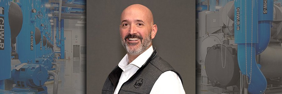 A man with a bald head and beard, wearing a white shirt and gray vest, is smiling at the camera. The background features industrial equipment and blue machinery.