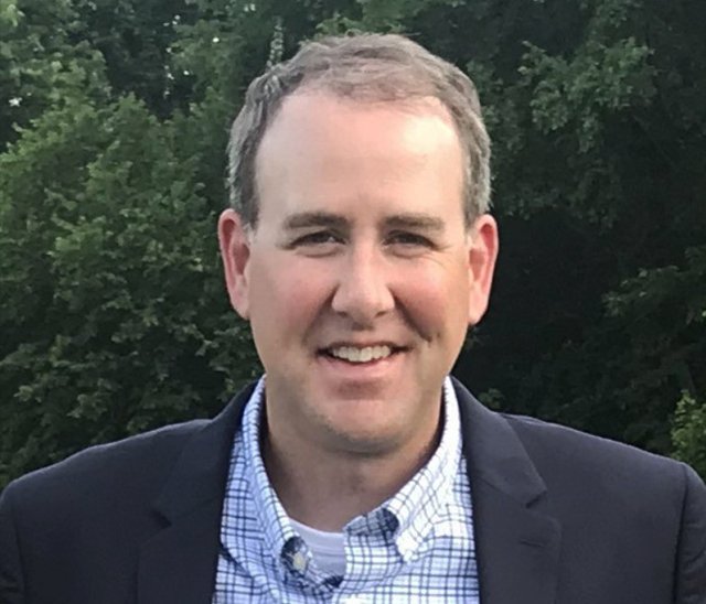 A man with short brown hair is wearing a navy blazer over a blue and white checked shirt. He is smiling and standing outdoors in front of green trees.