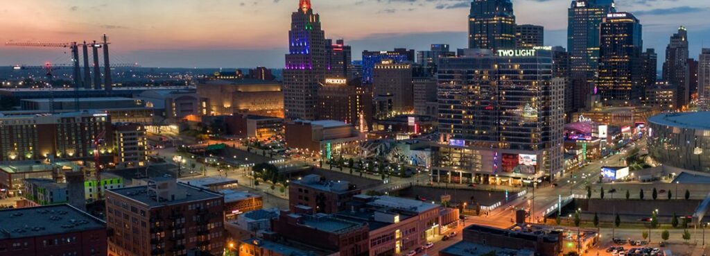 A cityscape of downtown Kansas City at dusk, showing tall buildings with lights on, including the Power & Light District, and visible streets with cars and pedestrians. The sky has a gradient from blue to orange near the horizon.