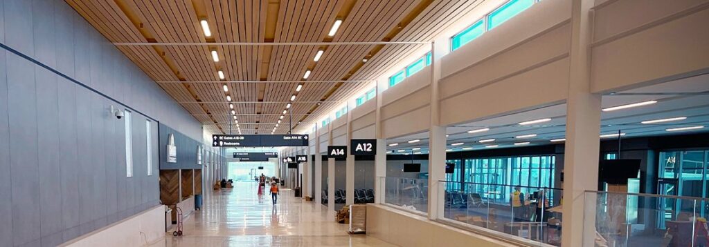A modern airport terminal with high ceilings, wide walkways, gate signs overhead, large windows on the right, and empty seating areas. Few people are visible in the distance near the gates.