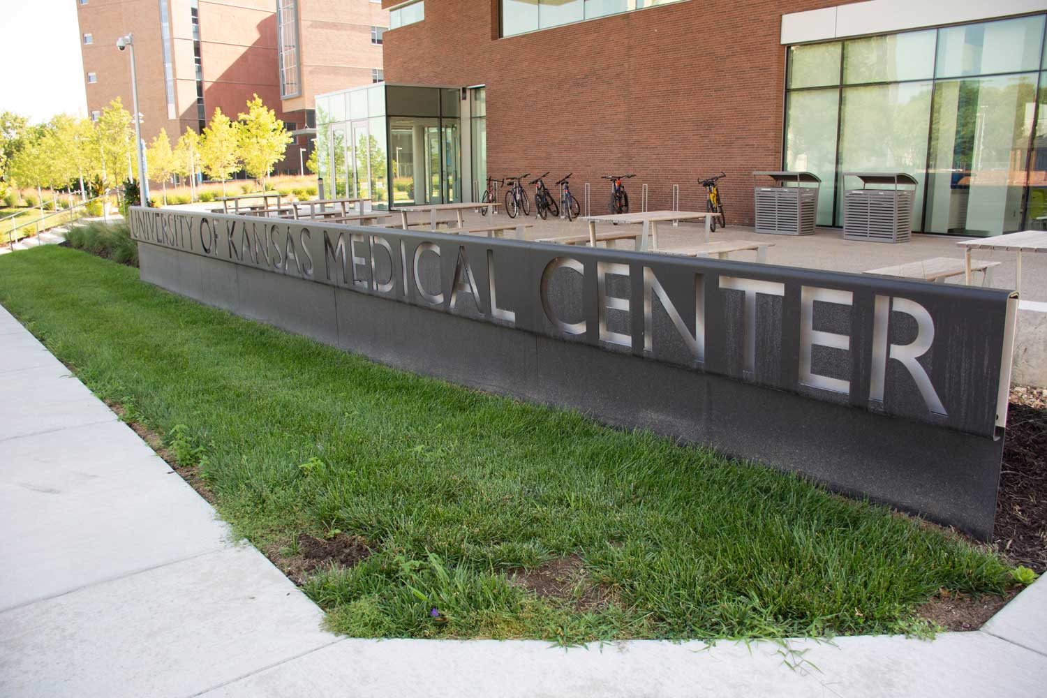 A large sign reading University of Kansas Medical Center stands outside a modern brick building with glass windows, bicycles, and green grass visible nearby.