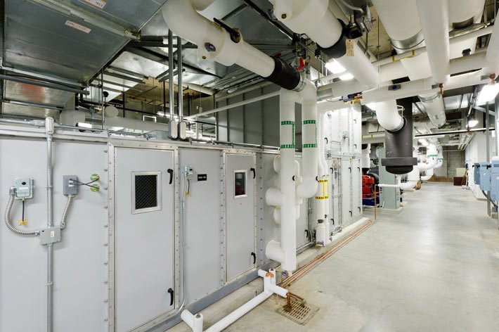 Industrial mechanical room with large HVAC units, exposed pipes, ventilation ducts, control panels, and a concrete floor. Overhead fluorescent lighting illuminates the equipment and structural elements.