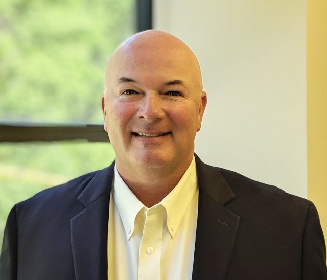 A middle-aged man with a bald head, wearing a dark suit jacket and white collared shirt, smiles at the camera. He is standing indoors in front of a window with greenery visible outside.