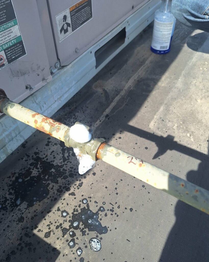 A soapy solution creates bubbles on a rusted metal pipe joint, likely indicating a leak test; nearby are some water stains, a safety warning label, and part of a spray bottle.