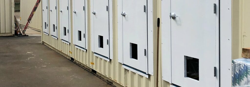 A row of beige metal structures with several white doors, each door featuring a small square window at the bottom. A ladder is visible in the background on the left side.