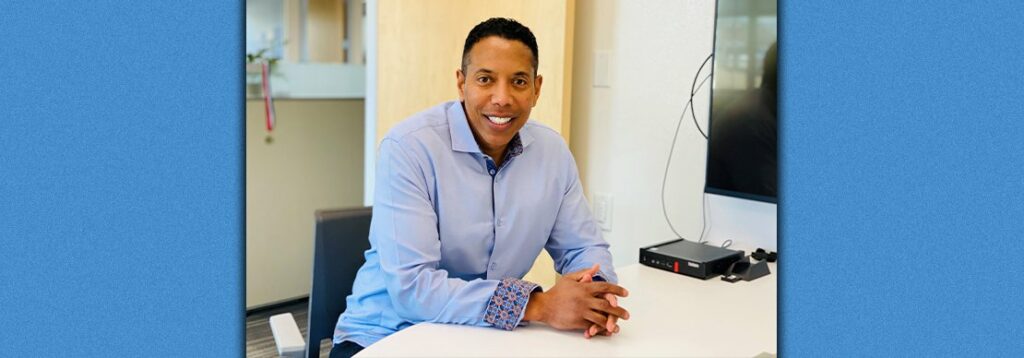 A man wearing a light blue dress shirt sits at a white table in an office, smiling at the camera. There is a television, a router, and cables on the wall beside him. The background shows an open door and office decor.