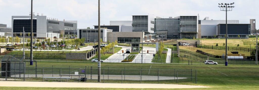 A large, modern building complex with glass and metal facades, surrounded by green lawns, parking areas, a baseball field in the foreground, and several cars on a road leading to the entrance.