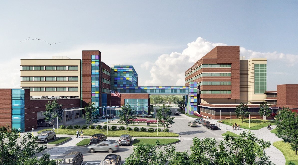 A modern hospital complex with multiple multi-story buildings, a glass skybridge, landscaped grounds, trees, parked cars, and people walking outside. An American flag is visible near the entrance.