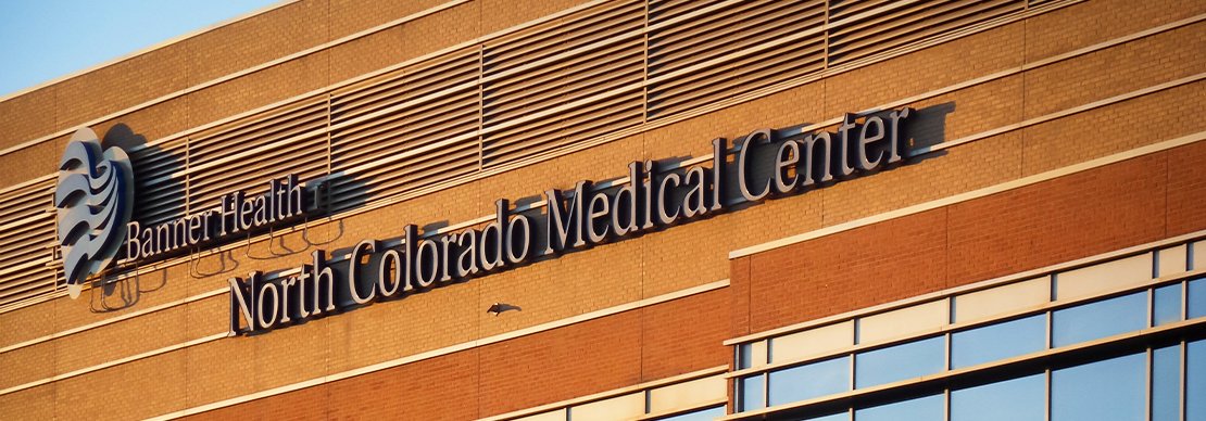 The exterior of a building with the sign Banner Health North Colorado Medical Center mounted on a brick wall. A bird is flying in front of the building.