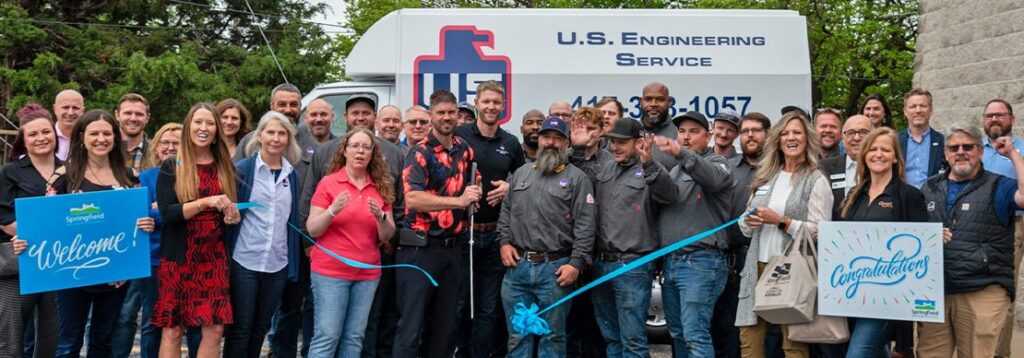 A group of people stand in front of a U.S. Engineering Service van, some holding a large blue ribbon and scissors, while others hold signs that say “Welcome” and “Congratulations.”.