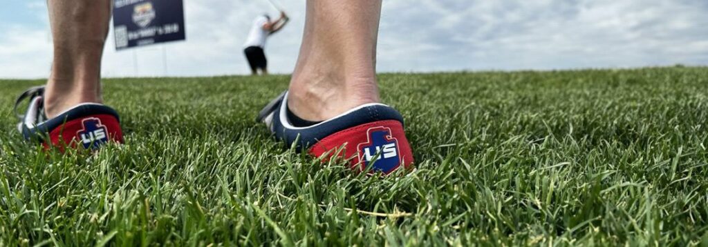 Close-up of a persons feet in blue and red shoes with US written on the heels, standing on grass. In the background, a person swings a golf club near a sign under a cloudy sky.